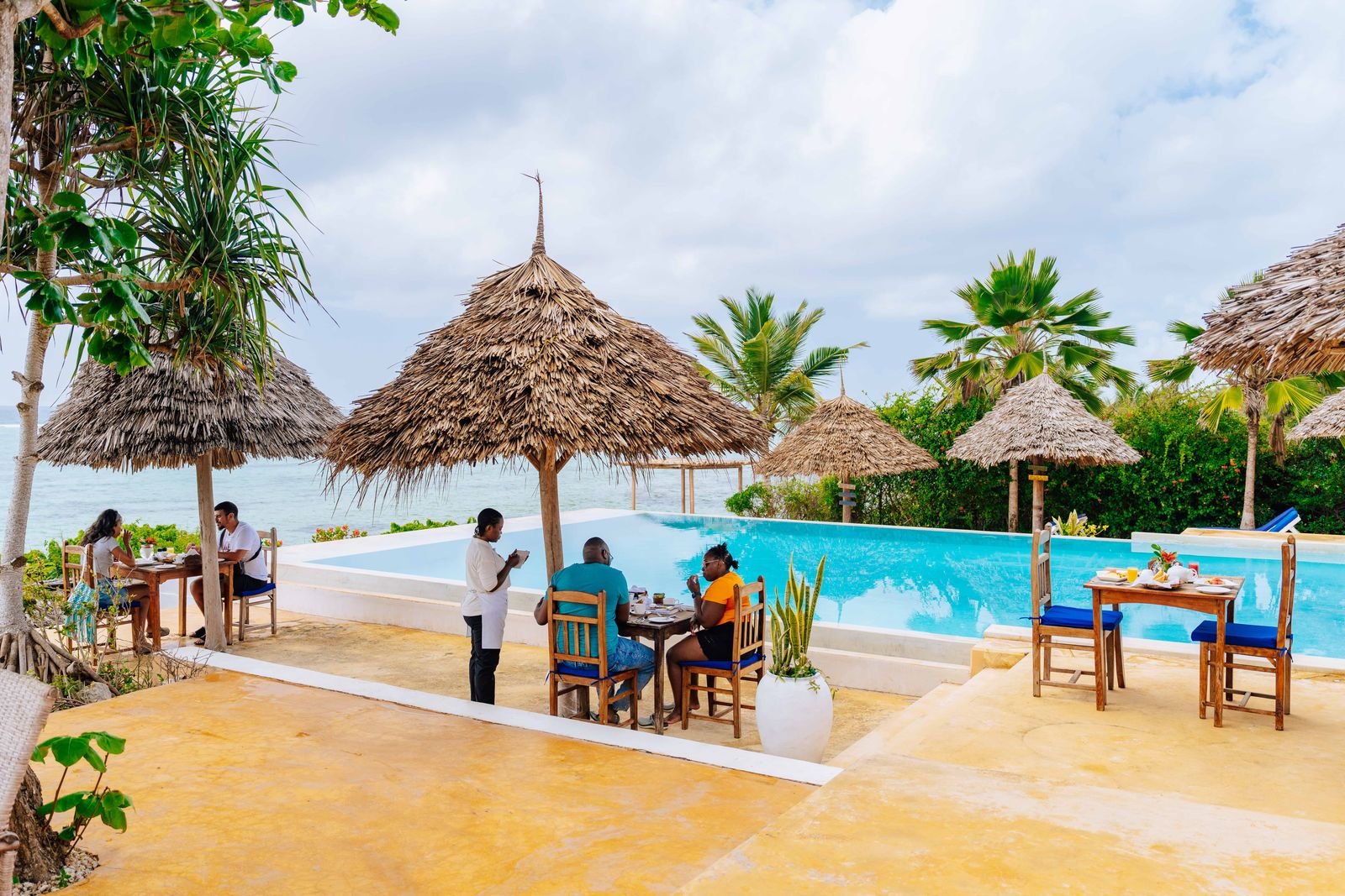 Guests dining by the pool under thatched umbrellas with the sea behind